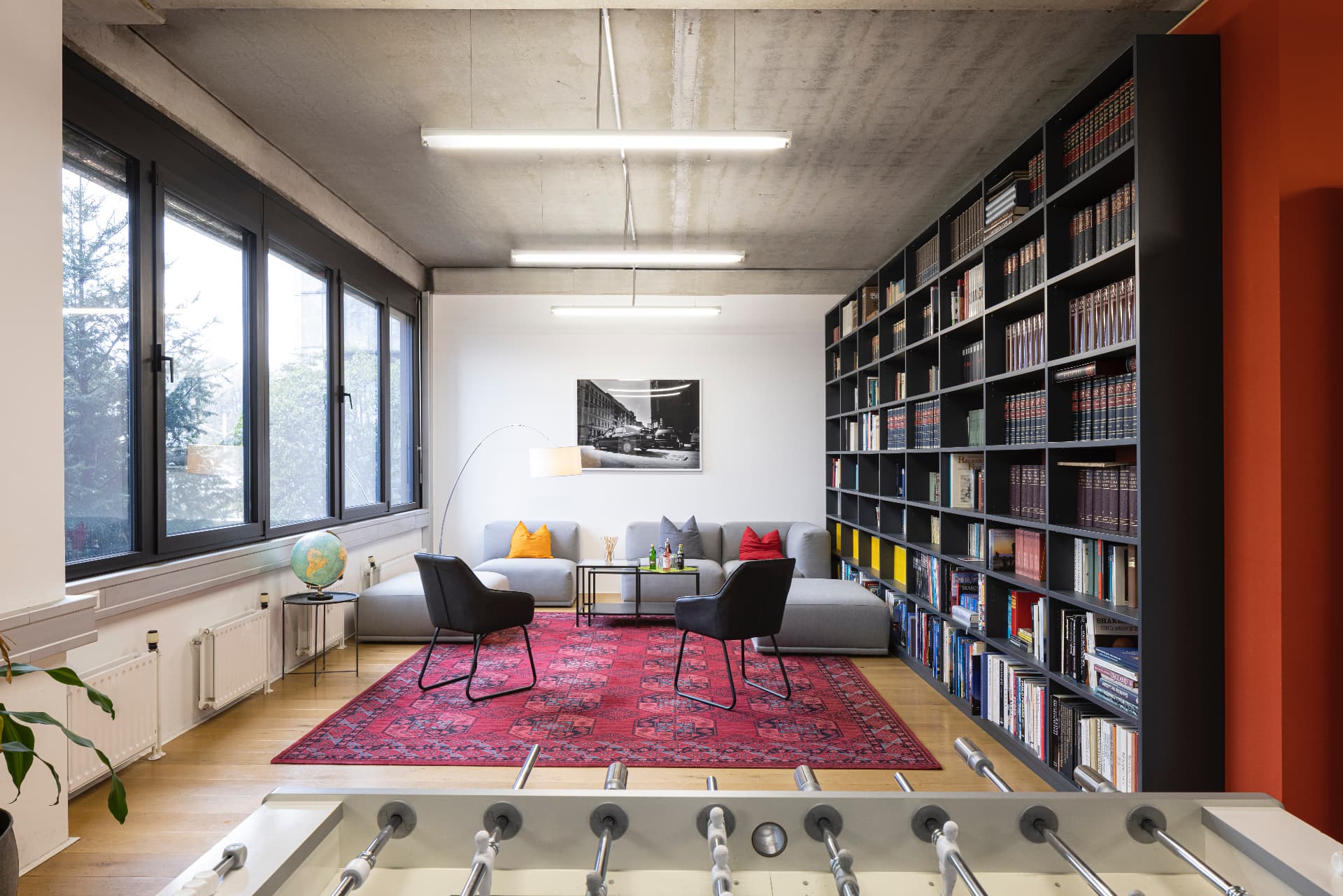 Library with a tall black bookshelf wall, grey sofa landscape, red rug, globe and a foosball table