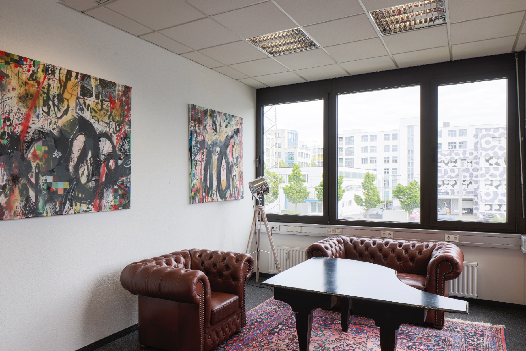 Two expressive large-format paintings above brown Chesterfield armchairs and a tripod studio lamp