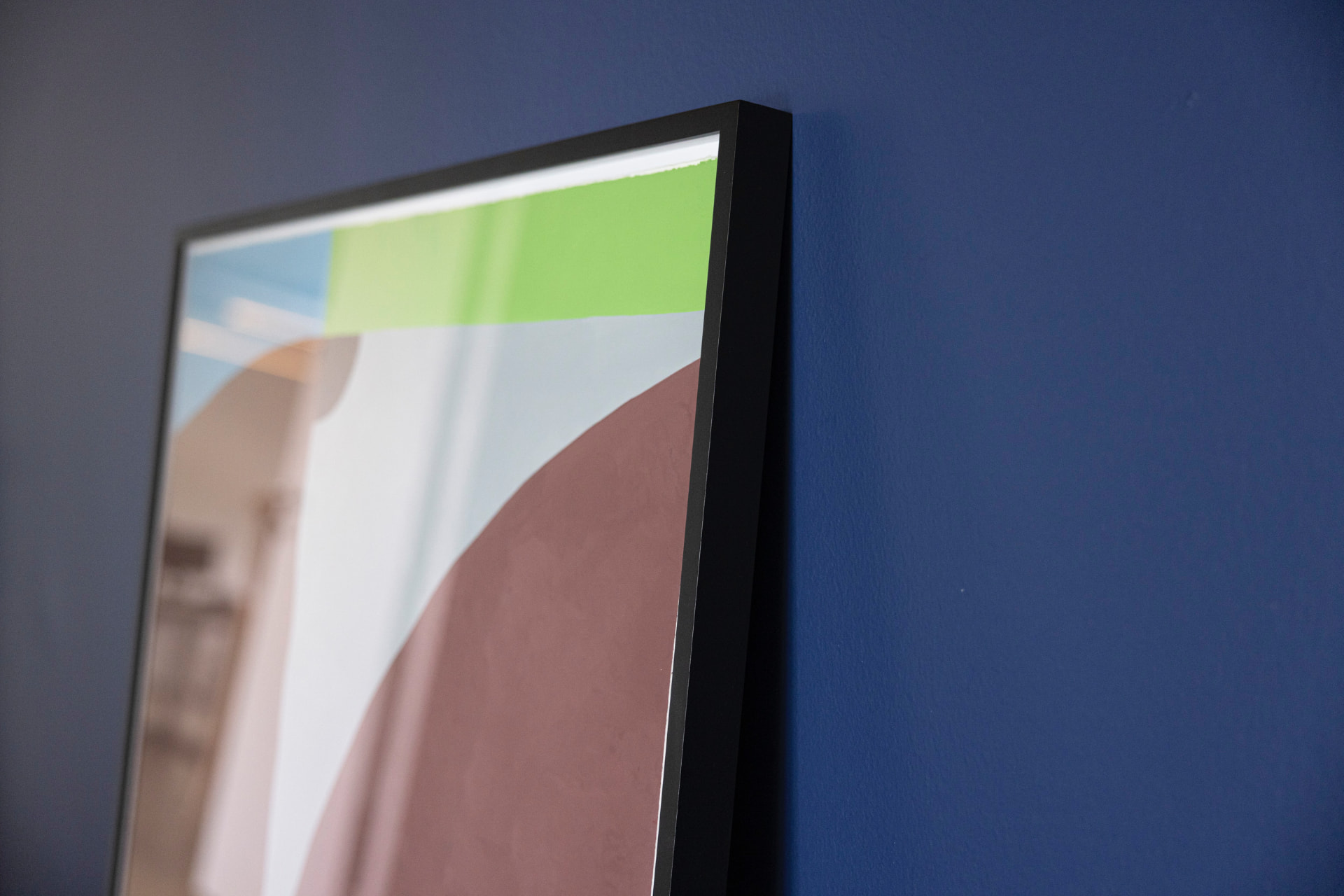 Close-up of a framed print with green, sky and mauve colour fields against a deep blue wall