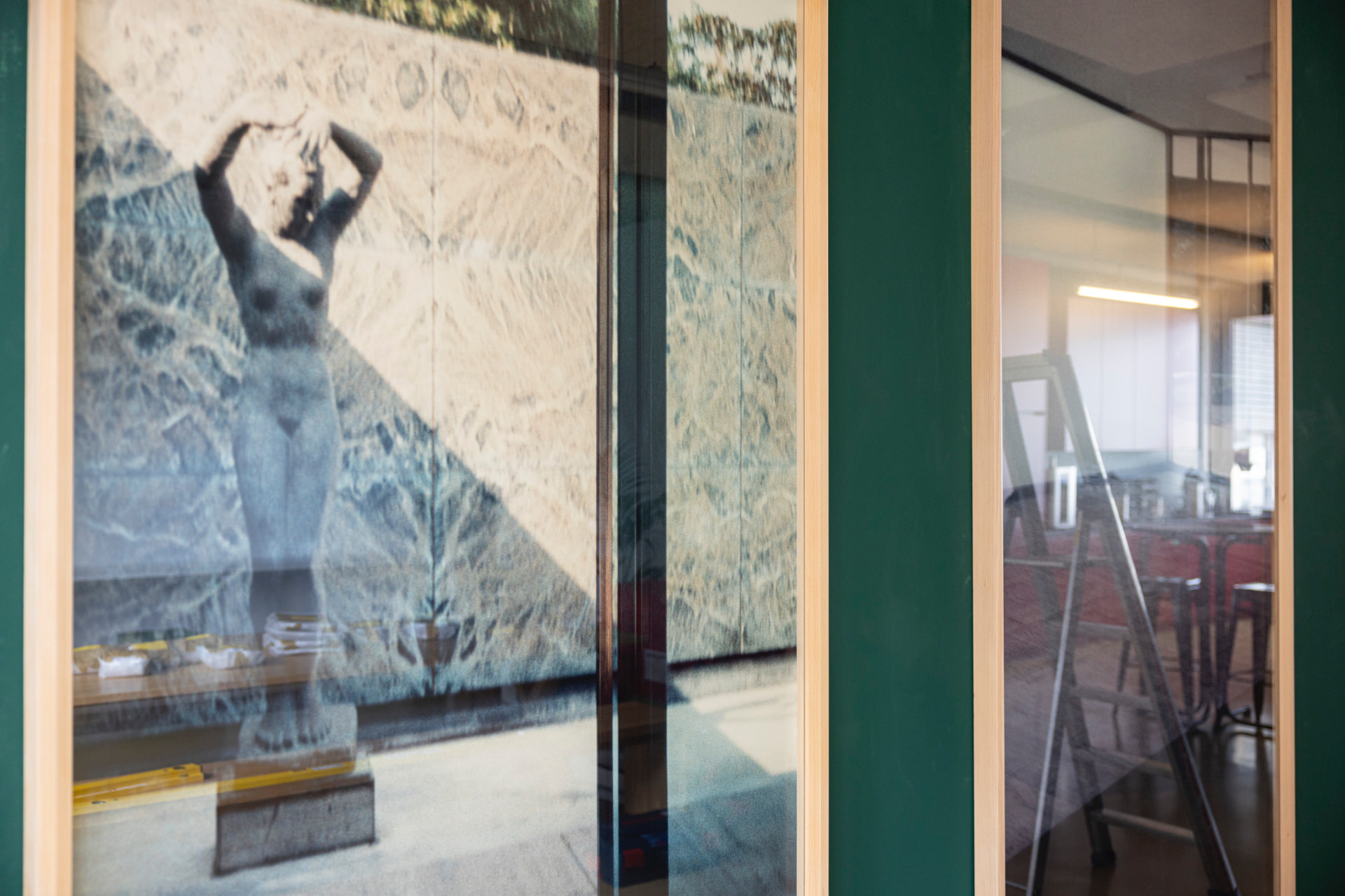 Framed photographic work of a nude figure sculpture behind glass, with green wall and reflections of the room