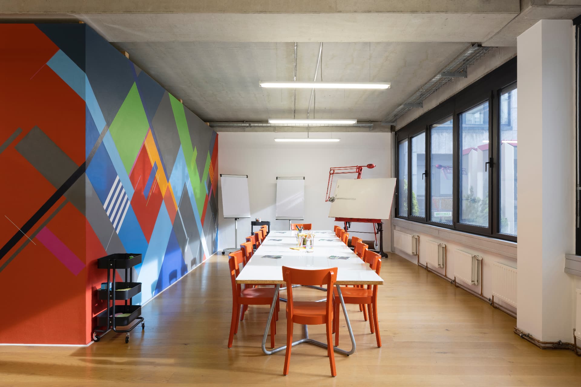 Workshop room with a large geometric wall painting in red, blue and green, orange chairs and a long white table